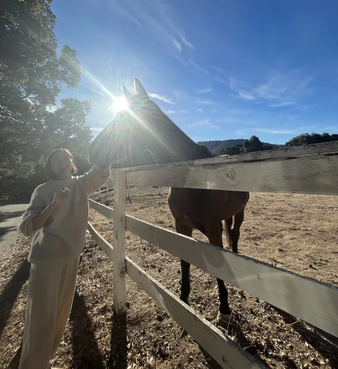 A person posing with a horse standing behind a fence. The sun is shining in the background.